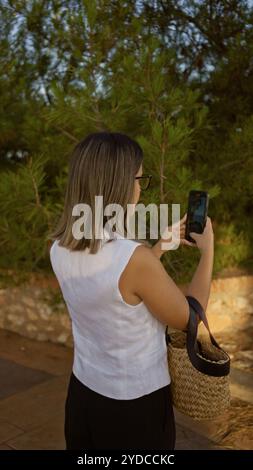 Junge hispanische Frau im Freien mit Smartphone und Brille, von hinten mit grünen Bäumen im Hintergrund an einem sonnigen Tag gefangen. Stockfoto