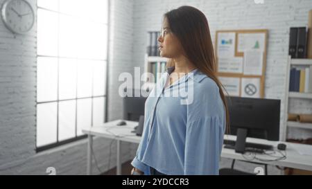 Junge Frau in einem hellen Bürozimmer mit Brille und blauer Bluse, die aus dem Fenster blickt, mit Computerarbeitsplätzen im Hintergrund Stockfoto