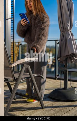 Frau auf einer sonnigen Terrasse im Herbst Prag, mit Kaffee und Smartphone in einem grauen Kunstpelzmantel Stockfoto