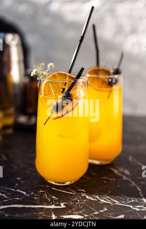 Nahaufnahme von zwei Gläsern mit einem Zitruscocktail auf einem schwarzen Marmortisch. Leckere gekühlte Getränke sind mit Orangenscheiben, getrockneten Blumen und Oliven dekoriert Stockfoto