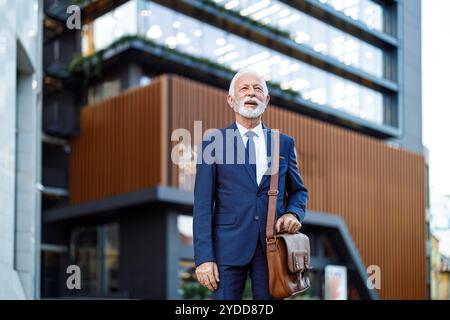 Senior männlicher Unternehmer mit Tasche, der wegblickt und nachdenkt, während er vor einem modernen Gebäude steht Stockfoto