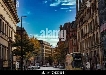 Eine Stadtstraße mit hohen Backsteinhäusern, einem gelben Doppeldeckerbus und Autos, die am Straßenrand geparkt sind. Der Himmel ist blau mit weißen Wolken im Menschen Stockfoto