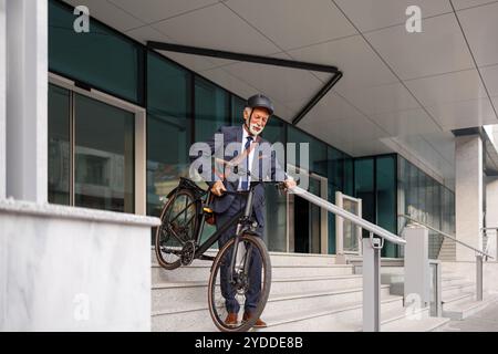 Die volle Länge eines bärtigen, älteren Unternehmers, der Fahrrad trägt und nach der Arbeit die Treppe hinuntergeht Stockfoto