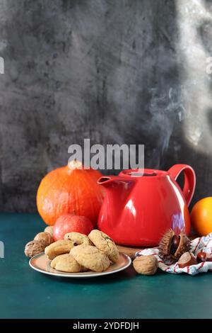 Rote dampfende Teekanne aus der Nähe. Heißer Tee in einem Topf und hausgemachte Kekse auf einem Tisch. Dunkler Hintergrund mit Kopierraum. Warmer Winterabend Stockfoto