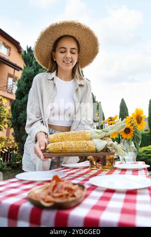 Schöne Frau mit frischen Maiskolben in der Nähe des Tisches im Garten Stockfoto