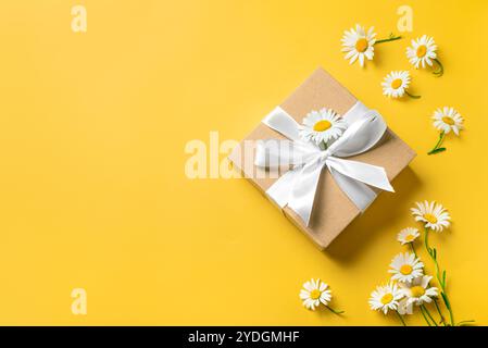 Geschenkbox mit weißem Band und Kamillenblüten auf hellgelbem Hintergrund, Blick von oben. Muttertag, Geburtstag, Jubiläumsfeier, Concep Stockfoto