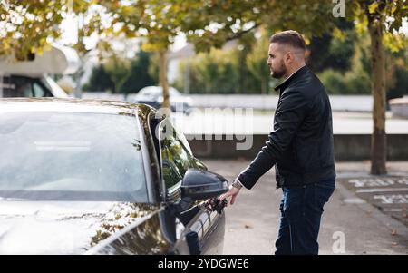 Gutaussehender bärtiger Kerl öffnet eine Tür seines Luxusautos auf dem Parkplatz. Der Mann beendet die Arbeit und fährt nach Hause Stockfoto