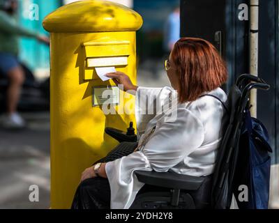 Frau im Rollstuhl postet einen Brief im gelben Briefkasten Stockfoto