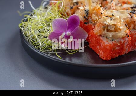 Nahaufnahme von lebendigem Sushi mit Bonitoflocken und schwarzem Sesam, garniert mit frischen violetten Orchideen und grünen Sprossen auf einem dunklen Teller Stockfoto