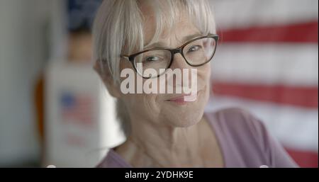 Große Nahaufnahme einer älteren Frau mit hochgezogenen Haaren, die vor die Kamera lächelt, während sie am Stand Stimmen mit US-Flagge hinter der Wand abgibt. Asiatischer Mann stimmt im Hintergrund ab Stockfoto