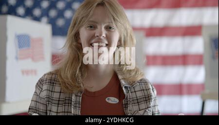 Eine junge Frau mit roten Haaren trägt einen „Ich habe gewählt“-Aufkleber und lächelt. Hinter ihr sind Wahlkabinen zu sehen, und die Flagge der Vereinigten Staaten auf der Rückwand im Hintergrund Stockfoto