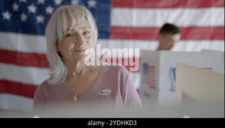 Ältere Wählerinnen tragen einen „Ich habe abgestimmt“-Aufkleber und lächeln vor die Kamera. Asiatische Wählerin, weicher Fokus, in der Kabine hinter ihr. US-Flagge an der Rückwand Stockfoto