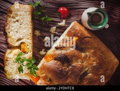 Rustikales Focaccia-Brot mit goldener Kruste, garniert mit Kräutern und Kirschtomaten. Präsentiert auf einer Holzoberfläche mit Petersilie, einer ganzen Kirschtomate, Stockfoto