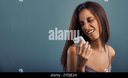 Die junge Frau ist vor Ekel grimmig, während sie eine Sprachnachricht auf ihrem Smartphone vor blauem Hintergrund aufnimmt und negative Gefühle ausdrückt Stockfoto
