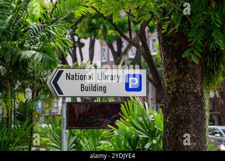 Ein großes Schild, das auf das Gebäude der Nationalbibliothek und den Parkplatz in Singapur zeigt, ist von Straßenbäumen und typischem Grün Singapurs umgeben Stockfoto