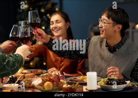Fröhlicher asiatischer Mann, der die Gläser anklickt, um Weihnachten mit Freunden zu feiern, die beim Beisammensein am Esstisch mit festlichen Gerichten anstoßen Stockfoto