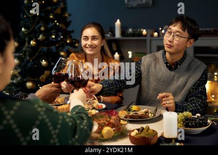 Mittelgroße Aufnahme eines asiatischen Mannes, der am festlichen Esstisch sitzt und Freunde die Gläser anklirrt, während er Weihnachten zu Hause feiert Stockfoto