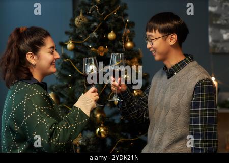 Seitenansicht eines fröhlichen, multiethnischen Paares, das Gläser mit Rotwein hält, während er den Feiertag feiert und im Wohnzimmer vor dem Weihnachtsbaum steht Stockfoto