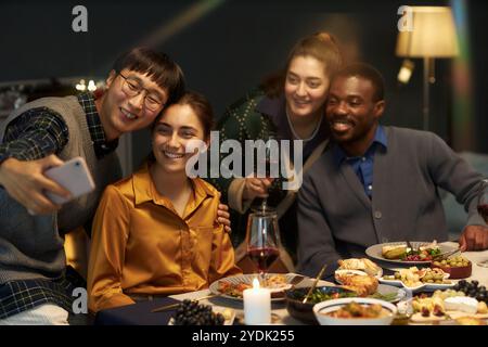 Freimütige Aufnahme eines asiatischen Mannes, der mit vier fröhlichen Freunden Selfie macht, während er am festlichen Esstisch sitzt, Weihnachten zu Hause feiert, Kopierraum Stockfoto