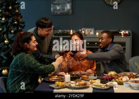 Verschiedene Gruppe von vier fröhlichen Freunden, die Weingläser anstoßen, um Weihnachten zu feiern, während sie am Esstisch sitzen, mit festlichen Gerichten zu Hause Dinner, Kopierraum Stockfoto
