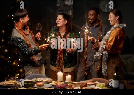 Mittelgroße Aufnahme von fröhlichen multiethnischen Freunden, die ein Glas Wein und leuchtende Glitzer halten, während sie im Wohnzimmer zu Hause miteinander plaudern und Weihnachten feiern Stockfoto