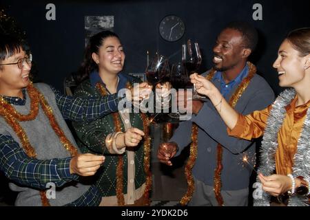 Mittelgroße Aufnahme einer Gruppe fröhlicher Freunde in Vintage-Outfits und Lametta, die Gläser mit Wein anklitzen, während sie gemeinsam Weihnachten im Wohnzimmer zu Hause feiern, Kamerablitz Stockfoto