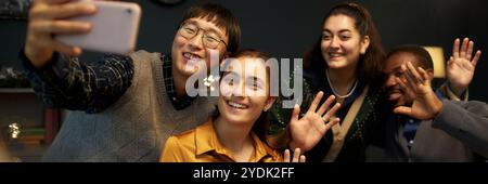 Freimütige Aufnahme eines asiatischen Mannes, der ein Selfie mit vier fröhlichen Freunden macht, die Hände vor der Kamera winken, während er am festlichen Esstisch sitzt und Weihnachten zu Hause feiert Stockfoto