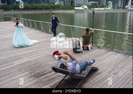 10.05.2019, Singapur, Republik Singapur, Asien, Ein Mann entspannt sich auf einer Liege an der Uferpromenade in Marina Bay, während ein Fotograf Hochzeitsfotos macht Stockfoto