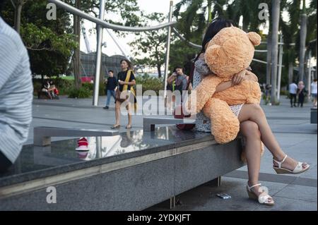 15.03.2019, Singapur, Republik Singapur, Asien, Eine junge Frau sitzt auf einer Bank am Ufer der Marina Bay und umarmt einen Teddybären, Asien Stockfoto