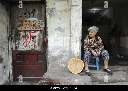 02.08.2012, Chongqing, China, Asien, eine alte Frau sitzt auf den Stufen vor ihrem Haus in der Altstadt von Chongqing. Gebürsteter Schriftzug an der Tür Stockfoto