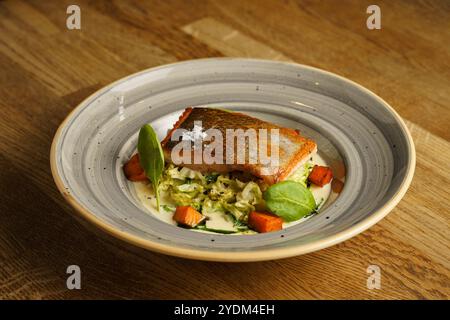 Ein perfekt gegrilltes Lachsfilet, serviert mit geröstetem Gemüse und einer cremigen Sauce auf einem dekorativen Teller. Das Gericht ist mit Spinatblättern garniert, Stockfoto