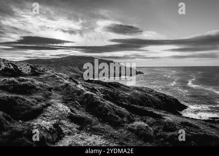 Die Umgebung bietet eine beeindruckende schwarz-weiße Küstenlandschaft, die zerklüftete Klippen und das turbulente Meer unter einem dramatischen Himmel in der Abenddämmerung hervorhebt. Stockfoto