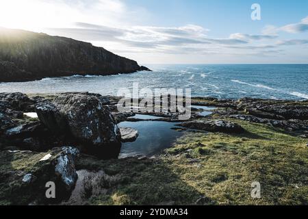 Eine malerische Küstenszene zeigt eine zerklüftete Küste mit großen Felsen, sanften Wellen und ruhigem Wasser unter einem klaren blauen Himmel bei Sonnenuntergang. Stockfoto