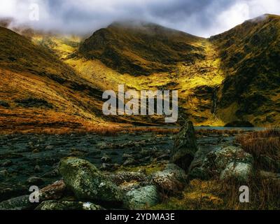 Ein kleiner, stiller See liegt zwischen zwei Berghängen, bedeckt mit schroffen Wolken und einer Verstreuung von großen, runden Felsen. Stockfoto