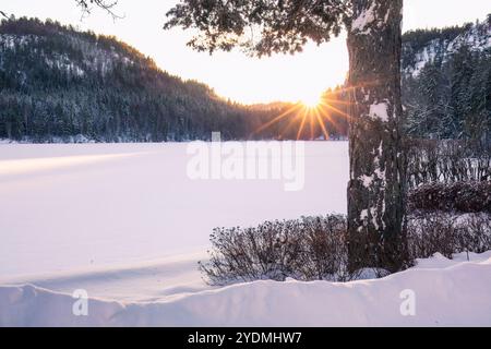 Ein heller Winteruntergang wirft warmes Licht über einen gefrorenen See und eine schneebedeckte Landschaft mit bewaldeten Hügeln im Hintergrund. Natürliche Schönheit, Winter Stockfoto