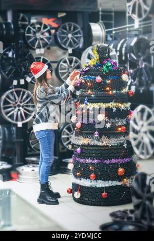 Ein Mädchen mit Weihnachtsmann-Hut schmückt kreativ einen Weihnachtsbaum aus Autoreifen mit Rädern um sie herum in einem Geschäft Stockfoto