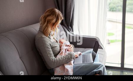 Berufstätige Mutter mit schlafendem Neugeborenen in den Armen, Multitasking mit Laptop, während sie zu Hause auf der Couch sitzt und Karriere und Mutterschaft in Einklang bringt Stockfoto