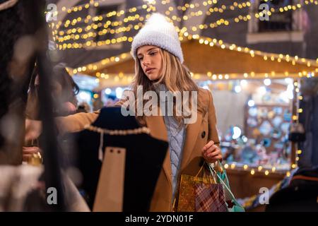 Wunderschöne junge Frau, die auf dem weihnachtsmarkt shoppt, festliche Stände unter Weihnachtslichtern erkundet - modische Kundin in Kamelmantel und weiß Stockfoto