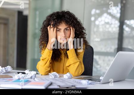 Gestresste lateinamerikanische Geschäftsfrau in gelbem Hemd fühlt Druck bei der Arbeit, umgeben von zerknitterten Papieren und Laptop. Konzept von Bürostress, Frustration, Überlastung. Stockfoto