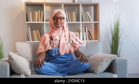 Arbeitsmeditation. Entspannender Arzt. Yoga Freizeit. Achtsame ruhige Frau Krankenschwester, die Stressentlastungsübungen in Lotusstellung auf der Couch im Krankenhaus praktiziert. Stockfoto