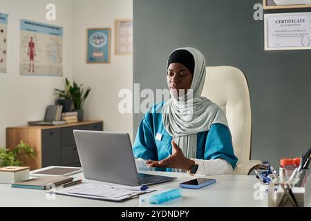 Fokussierte afroamerikanische muslimische Frau, die in der medizinischen Praxis arbeitet, Hijab und blaue Uniform trägt, am Schreibtisch sitzt mit Laptop und verschiedenen medizinischen Dokumenten ordentlich angeordnet Stockfoto