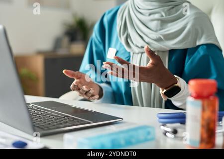 Muslimische Ärztin mit Gesten, während sie am Schreibtisch mit Laptop und Dokumenten sitzt, in einem Gespräch über die Arbeitsumgebung im Gesundheitswesen Stockfoto