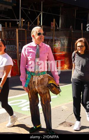 Ein stilvoller Mann mit Sonnenbrille, gestreiftem Hemd und einzigartiger Hose steht auf einer SOHO-Straße in Mann und hält einen Hut, während er mit zwei anderen Menschen geht Stockfoto