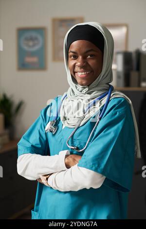Lächelnde muslimische Krankenschwester, die im Büro steht, mit Peelings und Stethoskopen, die Vertrauen und Wärme im Gesundheitswesen vermitteln Stockfoto
