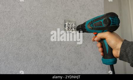 Ein Mitarbeiter, der eine Steckdose oder einen Schalter mit einem elektrischen Schraubendreher vor einem grauen Hintergrund repariert oder repariert Stockfoto