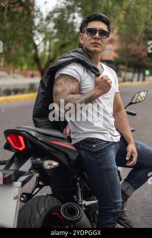 Attraktiver junger Mann mit kurzen Haaren, athletischer Körper, der auf seinem Motorrad sitzt, Brille trägt und seine Lederjacke hält, männlicher Model-Lifestyle Stockfoto