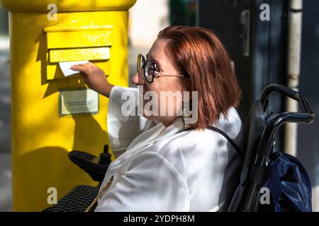 Lächelnde Frau im Rollstuhl, die einen Brief im gelben Briefkasten postet Stockfoto