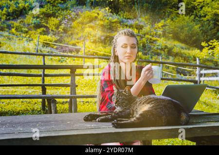 Eine junge Frau mit Dreadlocks, die ein rotes kariertes Hemd trägt, arbeitet draußen an einem Holztisch mit ihrer Katze und genießt eine Tasse Stockfoto