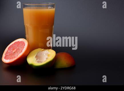 Eine Mischung aus frisch gepressten Säften aus tropischen Früchten auf schwarzem Hintergrund, neben den Hälften reifer Grapefruit, Mango und pitaya. Nahaufnahme. Stockfoto