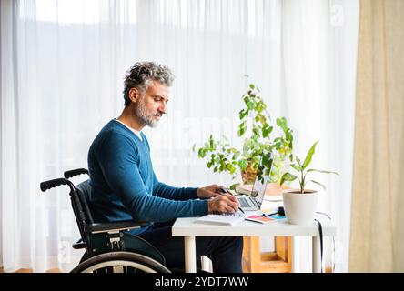 Mann mittleren Alters im Rollstuhl arbeitet vom Home Office aus, schreibt Notizen im Planer. Telearbeit und Home Office. Stockfoto